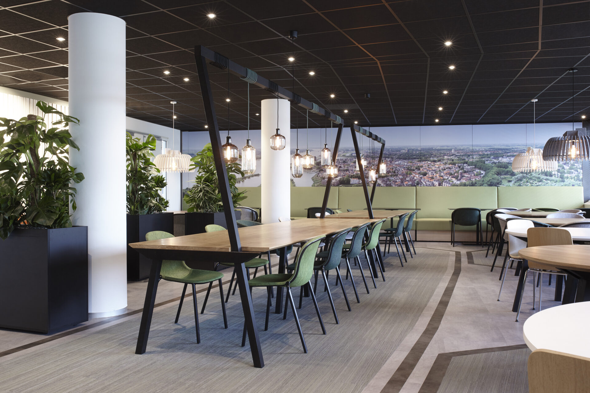 Bedrijfsrestaurant met lange houten tafels onder een zwart frame met hanglampen en groene stoelen