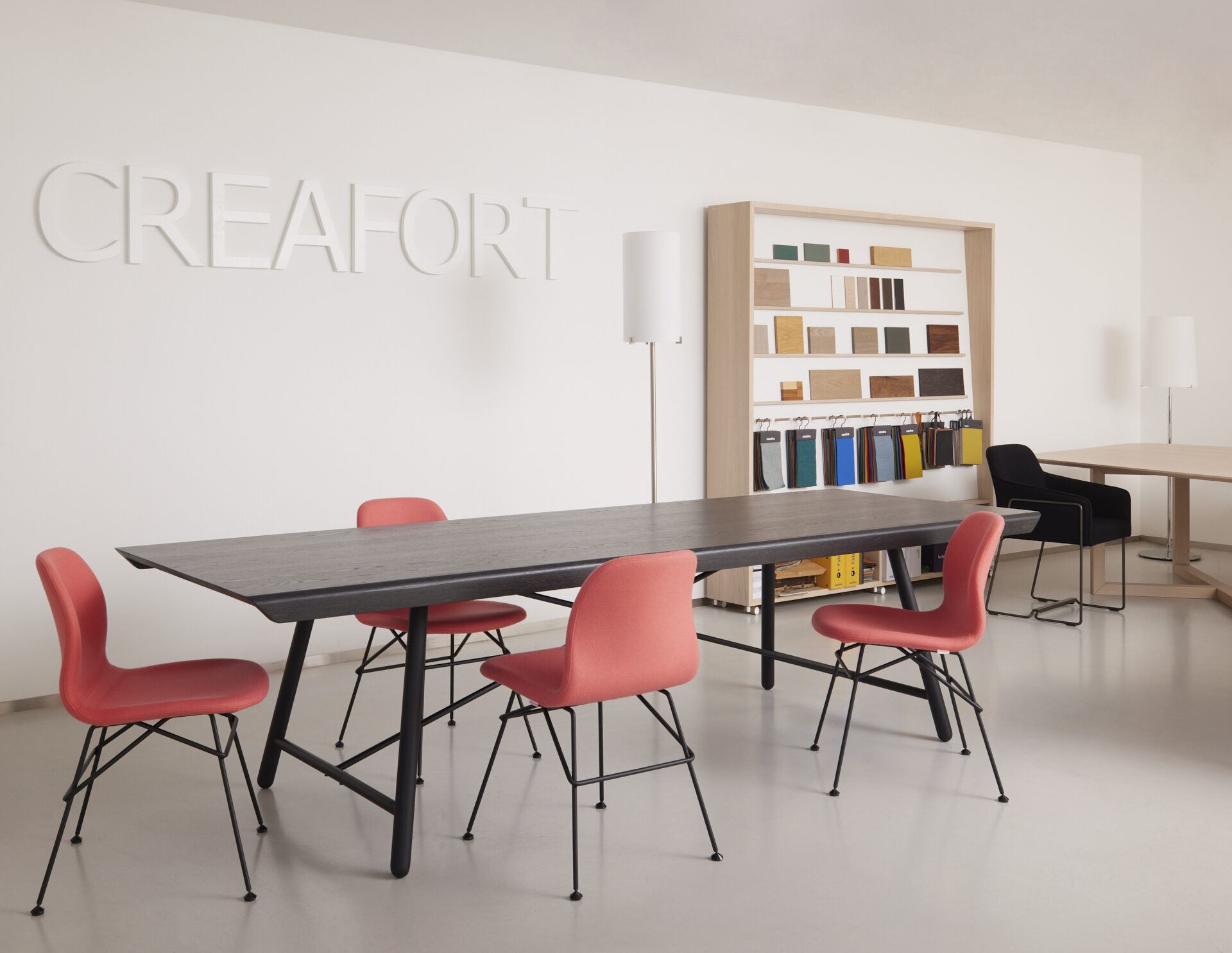 Ruimte met de wandbelettering Creafort, een wandrek met houtmonsters en stofstalen en een zwarte tafel met koraalrode stoelen