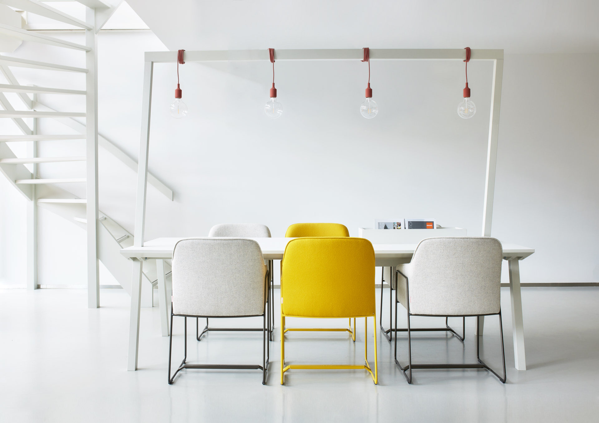 Witte tafel DRONE Lightning met een overspannend frame en vier hanglampen, met crème en gele fauteuils AVOLA in een witte ruimte met trap