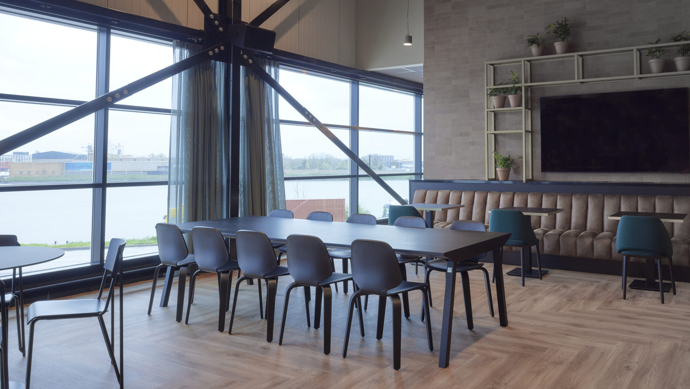Kantine met hoge glasgevel en uitzicht op het water, met een lange donkere tafel en zwarte stoelen