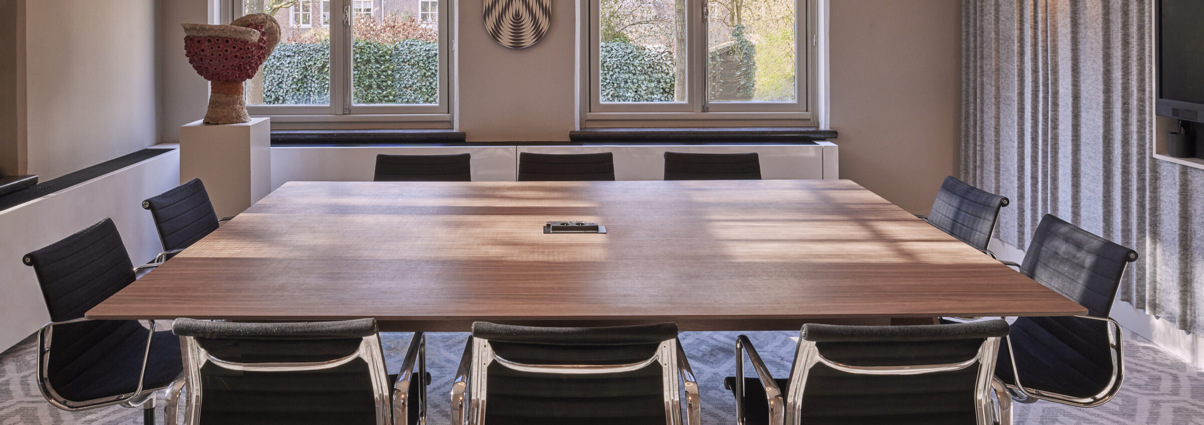 Vergaderkamer met een grote houten tafel en zwarte stoelen voor twee ramen met uitzicht op groen