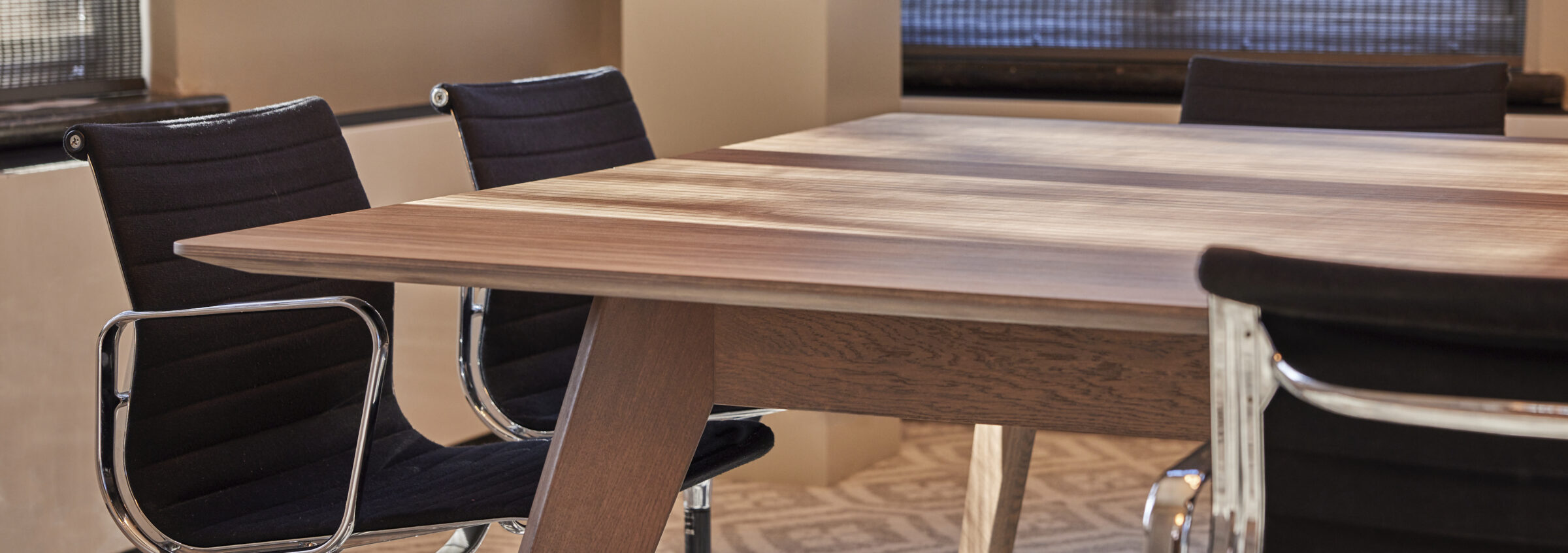 Hoek van een houten vergadertafel met schuine houten poot en afgeschuinde bladrand, met zwarte stoelen